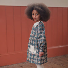 Load image into Gallery viewer, Young girl in a floral dress standing against a red paneled wall.