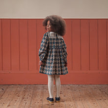 Load image into Gallery viewer, Child wearing a patterned dress standing in a room with wooden floor and red paneled walls.