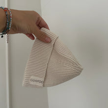 Load image into Gallery viewer, Hand holding a beige knit beanie with 'GUAPOO' branding against a white background