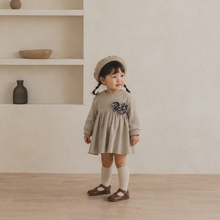 Load image into Gallery viewer, A toddler girl wearing a beige long-sleeve dress with a dark floral applique on the chest, a matching beige beret, white socks, and brown Mary Jane shoes, standing in a minimalist room with a textured cream wall and built-in shelves.
