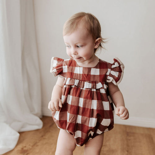 Baby wearing a red and white checkered romper standing on a wooden floor.