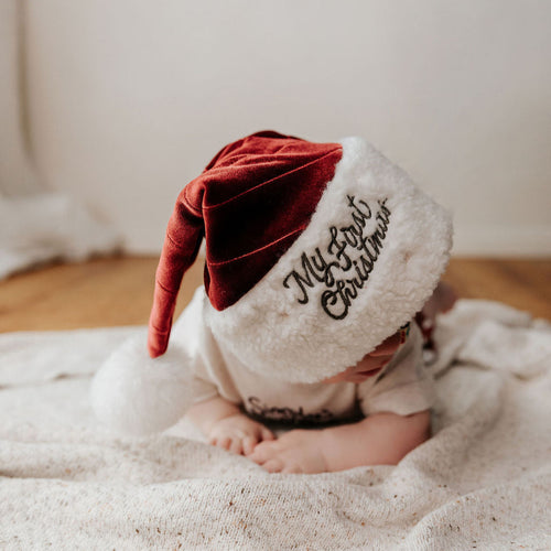 Baby wearing a white and red Christmas hat on a wooden floor.