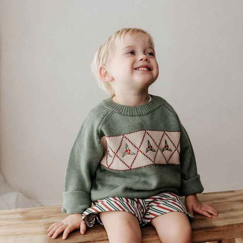 Child sitting on a wooden stool wearing a green sweater with a patterned design.
