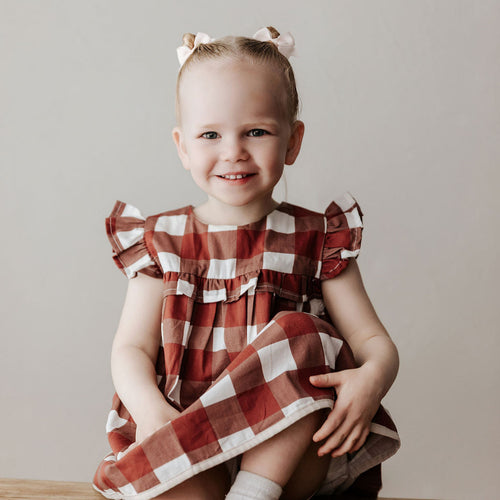 Young girl wearing a red and white checkered dress sitting on a wooden floor.