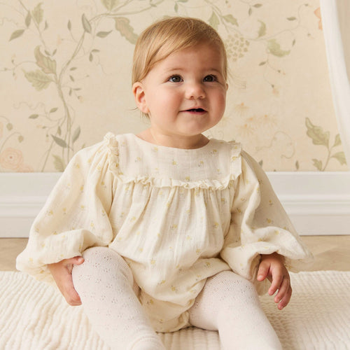 Baby in a white outfit sitting on a bed with floral wallpaper
