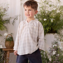 Load image into Gallery viewer, A young boy wearing a light beige and white plaid long-sleeve shirt with dark navy shorts and beige sandals, standing in a decorated indoor setting with plants and a wooden stool.
