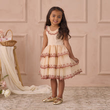 Load image into Gallery viewer, Young girl in a cream dress with red embroidery standing against a beige paneled wall.
