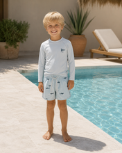 將圖片載入圖庫檢視器 A young blonde boy stands barefoot by a swimming pool wearing a light blue long-sleeve swim shirt with a small shark graphic on the chest and matching light blue swim shorts with a shark print.
