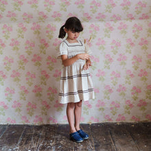Load image into Gallery viewer, Young girl in a white dress standing in a room with floral wallpaper.
